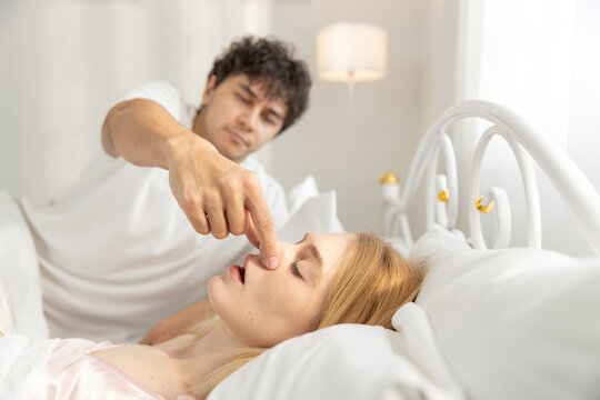 Frustrated man pinches women girlfriend nose to stop snoring while lying in bed. Concept of sleep disturbance, relationship issue, bedtime conflict, and noisy affecting rest quality.