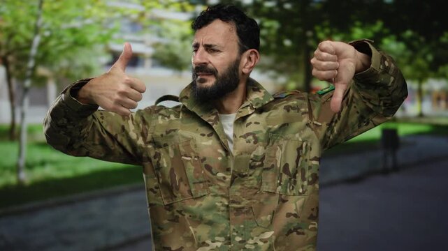 Hispanic military man with a beard in uniform making thumbs up and down gestures on a street outdoors, reflecting a contrast of opinions in an intriguing outdoor setting.