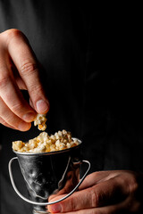 A person's hands hold a small metal bucket filled with freshly popped popcorn against a dark background, with a hand reaching in to take a piece.