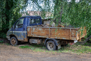 Rusty old blue pickup truck loaded with firewood, parked under leafy trees in a rural setting &mdash; perfect for themes of decay, nostalgia, countryside life, and industrial textures.