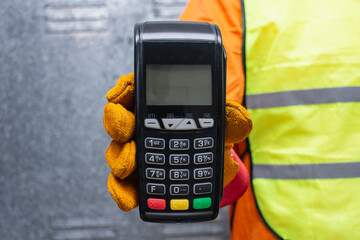 Construction worker wearing protective gloves and reflective workwear holding a wireless payment terminal. Concept of digital transactions, contactless payments in the construction industry.