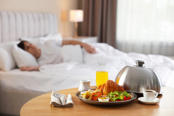 Delicious breakfast served on wooden table and man sleeping in bed at hotel room, selective focus