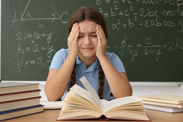 Back to school. Sad student with books at wooden desk against chalkboard with math assignment indoors