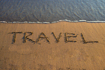 The word “TRAVEL” written on sandy beach near the shoreline, with gentle sea waves approaching.