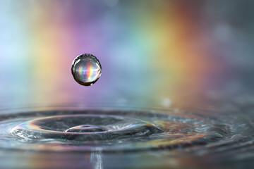 A water droplet is captured mid-air just above the surface, creating a stunning ripple effect with an iridescent rainbow of colors reflected from the surrounding light.