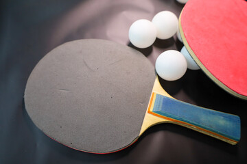 A close-up of two table tennis rackets and white balls scattered on a dark background. One paddle...