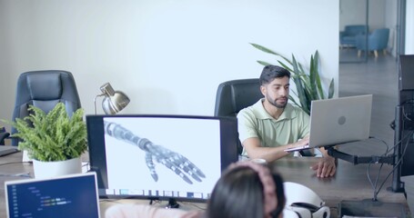 Working Indian man typing on laptop at open office desk, with robotic hand schematic, copy space