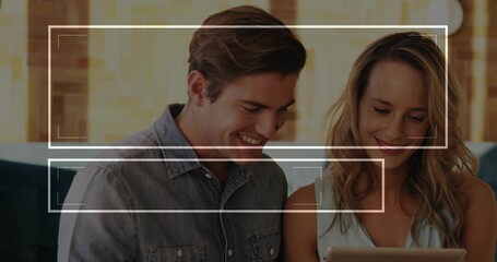 Smiling couple in denim shirt and sleeveless top leaning on sofa in living room, viewing tablet