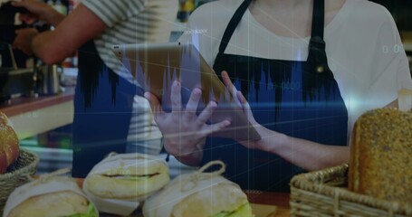Holding tablet, employee serving bagels and sandwiches at cafe counter, with baskets, portafilter