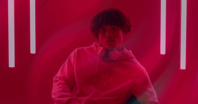 Sitting man wearing hoodie and over-ear headphones on chair in red studio, with vertical neon tubes - Powered by Adobe