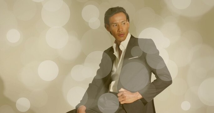 Posing adult Asian man on stool adjusting suit jacket and dress shirt in studio, bokeh backdrop - Powered by Adobe