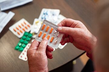 Elderly hands holding a blister pack of orange pills over a table with medications. Elderly hands holding pills, the daily struggle for health, dependence on medicine, the inevitability of aging