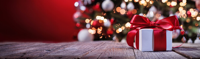 Gift with a Red Bow and a Christmas Tree. Wooden Table. Red Background
