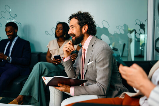 Smiling Caucasian businessman actively participates in a conversation while holding his planner with diverse male and female colleagues in a modern office lounge