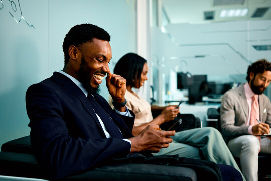 Smiling African American businessman in a suit laughs while looking at his mobile phone next to diverse colleagues in a modern office waiting area