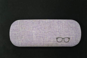 Close-up of a lilac, light purple hard eyeglasses case with a linen texture and a simple glasses icon, placed on a dark black background. Represents eye care, fashion, and product safety.
