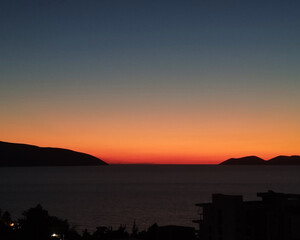 Vibrant orange sunset over the sea with silhouetted mountains and buildings in the foreground.