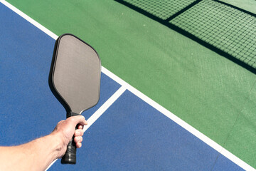 POV view of hand holding a pickleball paddle with a green and blue outdoor court surface in the background