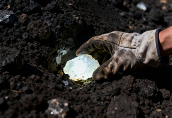 Obraz premium Gloved hand retrieving a glowing, translucent crystal or quartz from dark, rocky soil during excavation.