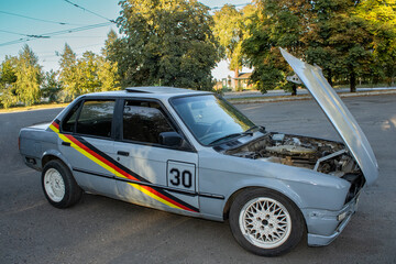 Old gray race car with open hood parked on street near trees