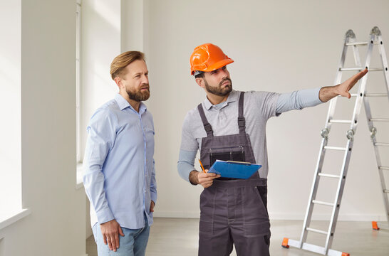 Construction worker in uniform and hard hat discusses renovation details with male client in modern apartment. They stand near a ladder, examining interior space during remodeling consultation.