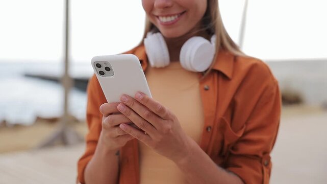 Close up young woman wears orange shirt casual clothes headphones hold use mobile cell phone rest relax in free time walk on sea ocean sand shore beach outdoor seaside in summer day. Lifestyle concept