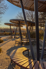 A sunlit park pavilion with curved wooden benches supported by tall metal columns. Outdoor modern seating area and visitors stroll through the public park landscape.