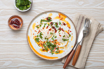 Turkish eggs cilbir, a delicious and colorful breakfast with poached eggs on yogurt, seasoned with spices and fresh herbs, arranged on a light wooden table