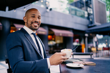 Cheerful businessman with wide smile holding coffee and smartphone, sitting outdoors, expressing confidence, digital comfort and a balanced mobile work atmosphere.