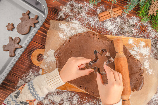 Hands cutting gingerbread dough with cookie cutter on wooden board, Christmas baking process with flour, rolling pin, cinnamon sticks, fir branches, and baking tray, cozy holiday atmosphere, top view. - Powered by Adobe