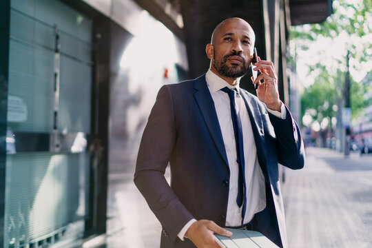 Confident businessman walking while talking on smartphone and holding tablet, portraying mobile communication, multitasking and productivity in urban tech-driven business routine.