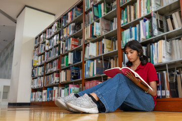 Teenage student girl sitting on the library floor, reading book beside bookshelves in educational school.
