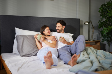 Fototapeta premium A couple relaxes in a modern bedroom, smiling at each other while holding mugs. Soft bedding and natural light create a warm, inviting atmosphere for their morning together.
