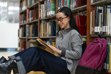 Young woman student sitting on the library floor near bookshelf, smiling while reading book in college library. learning or information in university.