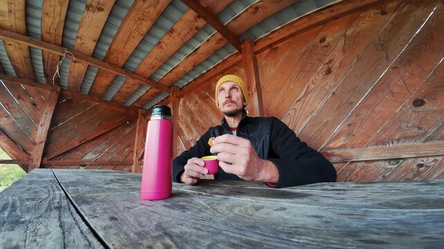 A man enjoys a warm drink and snack in a cozy rustic shelter, surrounded by beautiful naturea perfect setting for outdoor enthusiasts and lifestyle content creators to find inspiration