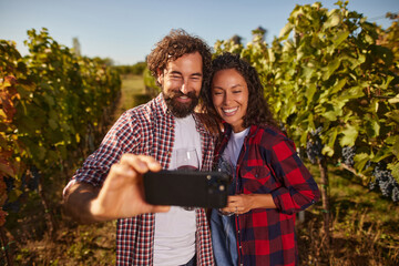 A couple smiles and holds a phone for a selfie amid lush vines in a vineyard. They celebrate their day with wine and laughter, surrounded by ripe grapes.