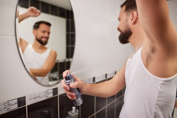 Obraz premium A man stands in front of a mirror, applying deodorant in a bathroom. He wears a tank top and smiles as he gets ready for his day.