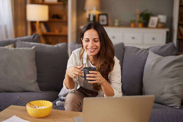 A relaxed woman smiles as she holds a coffee cup while sitting on a couch. A bowl of snacks is on the table beside her laptop, creating a cozy atmosphere in the living room.