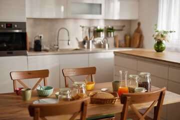 A family gathers around a wooden table in a sunny kitchen, sharing snacks and drinks. Colorful bowls and jars are laid out, creating a warm and inviting atmosphere.