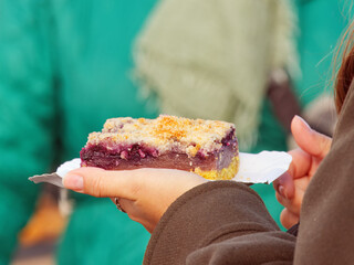 Delicate slice of homemade fruit cake with crumb topping, held in hand on a sunny autumn day at Prague Naplavka market. A sweet moment full of color, taste, and warmth.