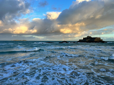 Golden sunlight illuminates waves and fortress under dramatic clouds, perfect for travel, tourism, and design use