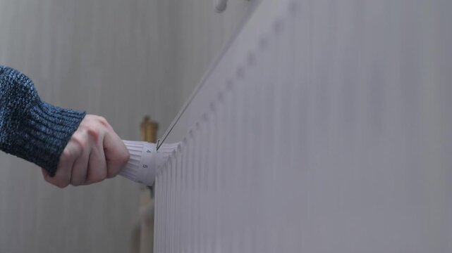 The hand of a man dressed in a sweater turns the heat regulator on a heating radiator in a house, the man is freezing inside the house and adjusts the heat of the heating batteries