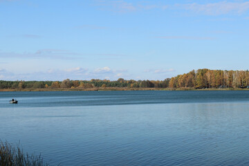 Panorama of a forest lake with a small fishing boat sailing in the distance, on a calm clear autumn day.