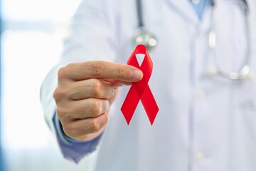 A male doctor in a white coat holds a red awareness ribbon. The background is blurred, suggesting a medical environment. The ribbon symbolizes health awareness.