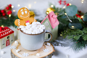 Gingerbread man cookie with marshmallows in mug on wooden slice surrounded by festive decor