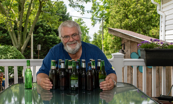 A cheerful senior man sits at a table on a sunny patio, surrounded by several bottles of beer. He smiles as he poses with his collection on a warm summer day, enjoying the outdoors.