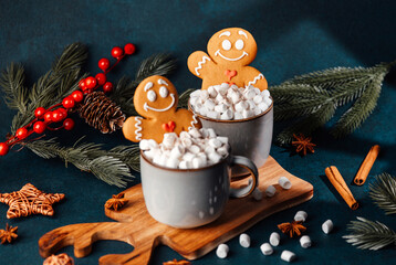 Two mugs with marshmallows and gingerbread men, festive winter drink, cinnamon sticks and pine branches.