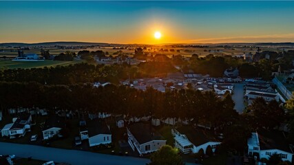 Golden sunlight breaks over the horizon, illuminating a quiet manufactured home residential area surrounded by lush fields. The morning mist adds a peaceful ambiance to the approaching day.