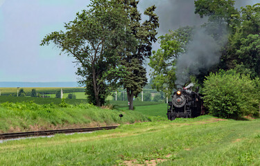 A vintage steam locomotive emits puffs of smoke as it moves along the tracks, surrounded by vibrant green fields and trees under a clear blue sky.