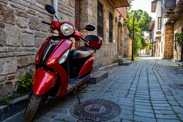 A glossy red scooter stands against a stone wall on a deserted cobblestone street in Kaleici,...